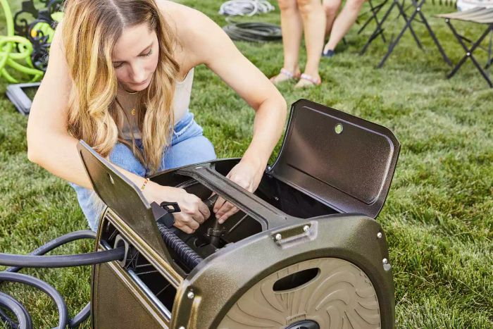 A woman attaching a hose to the Giraffe Metal Hose Reel Box on a grassy area