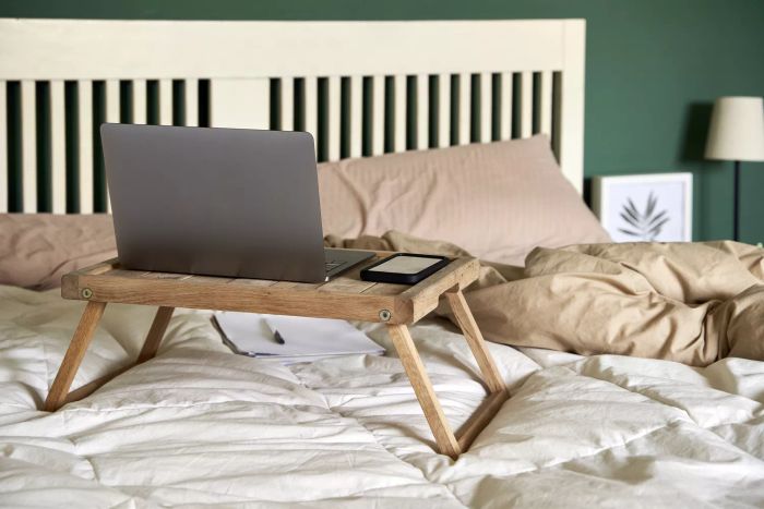 A wooden lap desk placed on a bed with a phone and laptop resting on it.