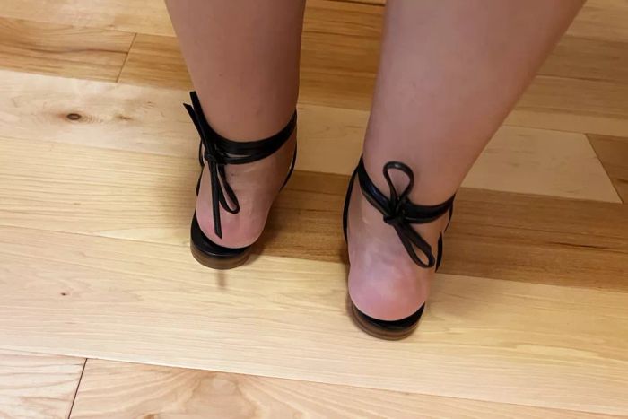 Heels of feet wearing Margaux The Wrap Sandals on a wooden floor