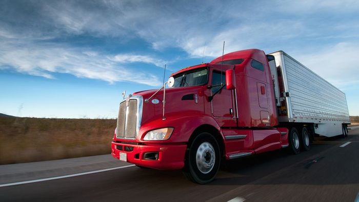A red semi truck hauling a white trailer cruises down the highway under the bright sun.