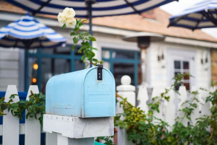 A traditional blue mailbox positioned in front of a house fence and a flower garden (blurred in the background). Close-up shot with selective focus.