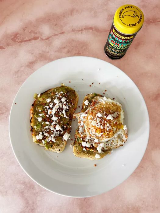 A plate of avocado toast topped with an egg and crumbled queso fresco, sitting next to a bottle of Trader Joe's Crunchy Chili Onion Sprinkle seasoning on a marble countertop.