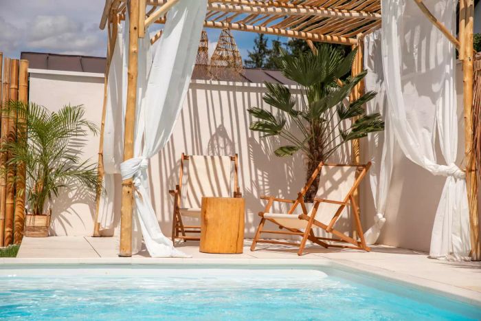 A relaxing Boho style with sun chairs placed under a driftwood pergola beside a shimmering pool, bathed in warm sunlight.