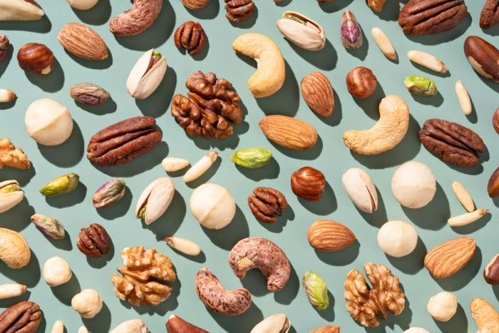A Variety of Nuts Including Almonds, Brazil Nuts, Cashews, Hazelnuts, Macadamia Nuts, Pecans, Pine Nuts, Pistachios, and Walnuts on a Flat Lay Green Background, Viewed from Above.