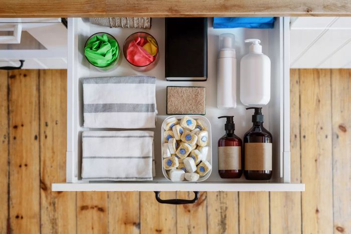 Aerial view of an open drawer containing towels, a washing sponge, detergent bottles, and dishwashing tablets neatly arranged in a plastic box inside a modern kitchen cabinet, showcasing effective storage solutions.