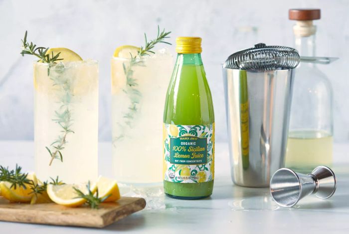 Trader Joe's Organic 100% Sicilian Lemon Juice is featured in the recipe for Sicilian Tom Collins, served in two tall glasses with garnishes of rosemary sprigs and lemon wedges.
