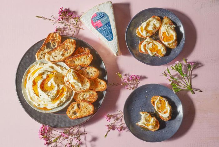 This is a photo of Trader Joe's Double Cream Brie, shown here as a whipped version topped with spicy honey sauce and cracked pepper, served on a sleek black plate with toasted crostini on the side.