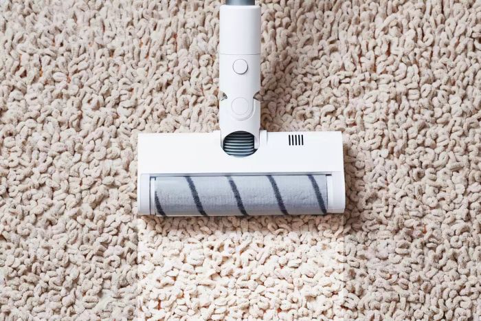 A person’s legs, accompanied by the white turbo brush of a cordless vacuum, effortlessly clean the carpet in a home. Advanced cleaning technology at work.