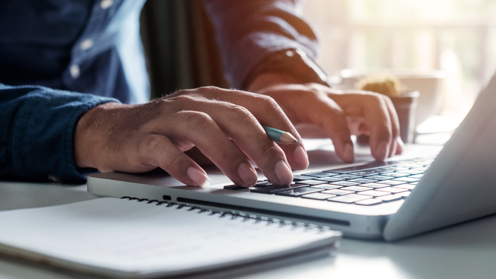A person typing on a laptop while holding a pencil in hand