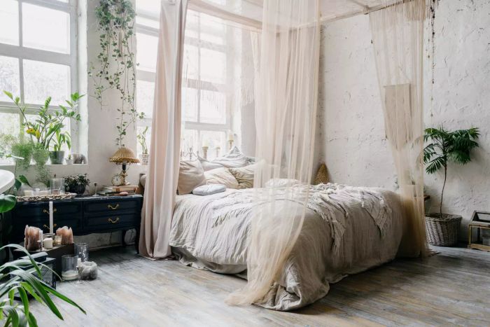 A bohemian-inspired cozy bedroom featuring a king-sized bed with a canopy, large windows on the wall behind it, and black antique furniture on the left side adorned with greenery, a vintage lamp, clock, and candles