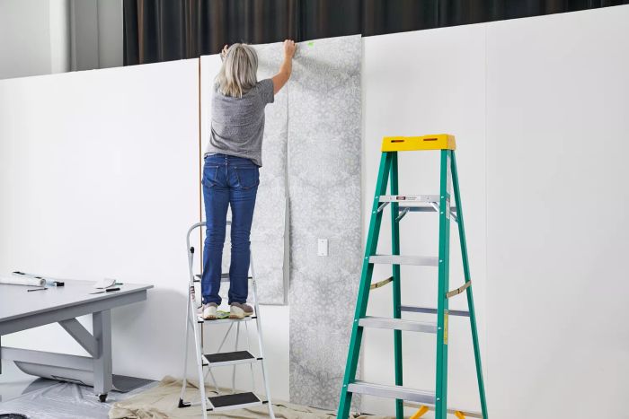 A person on a ladder applies the Ballard Designs Juliet Floral Wallpaper to a white wall.