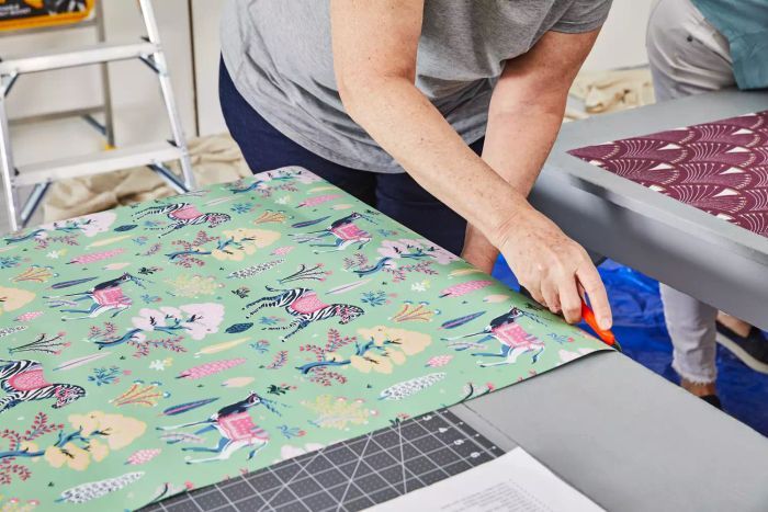A woman carefully cuts the Printfresh Antelope's Forest Wallpaper on a table with a snap blade