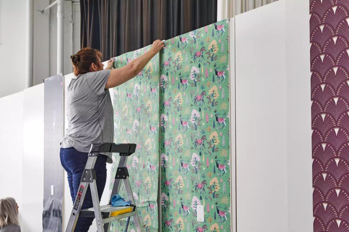 A person is standing on a ladder, applying the Printfresh Antelope's Forest Wallpaper to a white wall.
