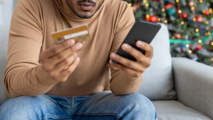 Frustrated man in a living room beside a decorated Christmas tree, checking his phone