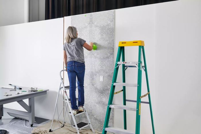 A person is standing on a ladder, applying the Ballard Designs Juliet Floral Wallpaper to a white wall using a sponge.