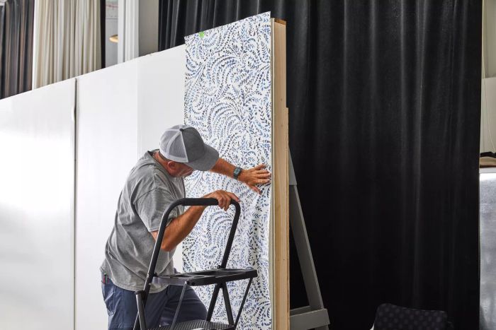 A person is seen standing on a ladder, applying the Serena & Lily Priano Wallpaper to a white surface.