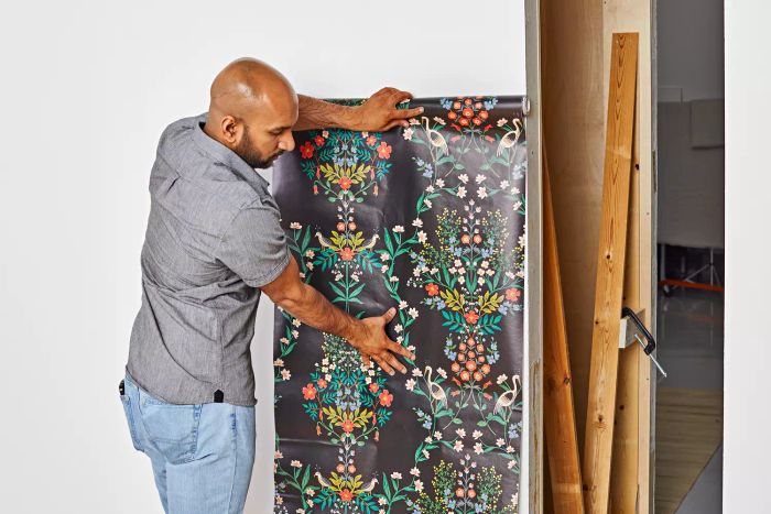 A person unrolls the Rifle Paper Co. Luxembourg Peel & Stick Wallpaper against a white wall, prepping for installation.