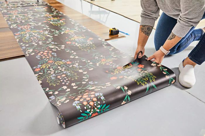 A person trims the Rifle Paper Co. Luxembourg Peel & Stick Wallpaper on the floor before applying it to the wall.
