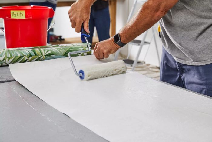 An individual applying adhesive using a paint roller on A-Street Prints Alfresco Palm Leaf Wallpaper