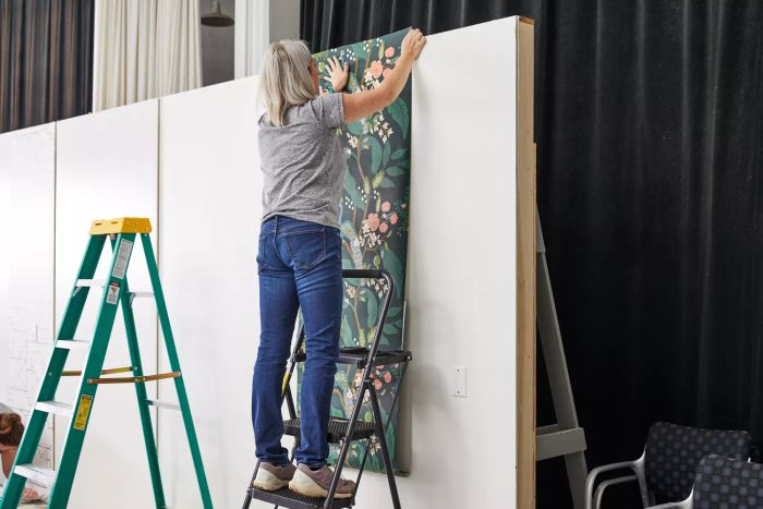 A woman applies Rifle Paper Co.'s Peacock Wallpaper to a white wall