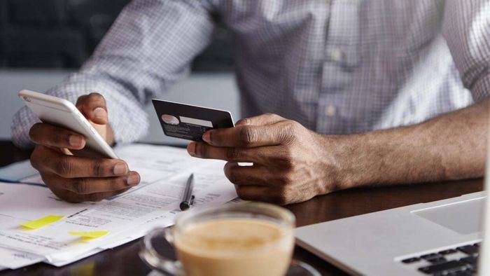 A man seated at a cluttered desk holds a credit card beside his smartphone