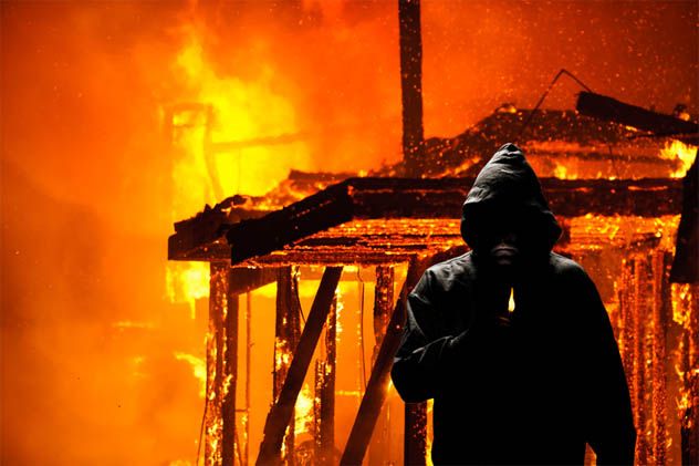 A hooded figure holding a lighter in front of a burning house