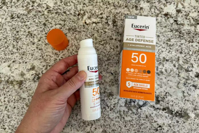 A hand holding an opened bottle of Eucerin Age Defense Face Sunscreen Tinted Lotion SPF 50 next to its opened box