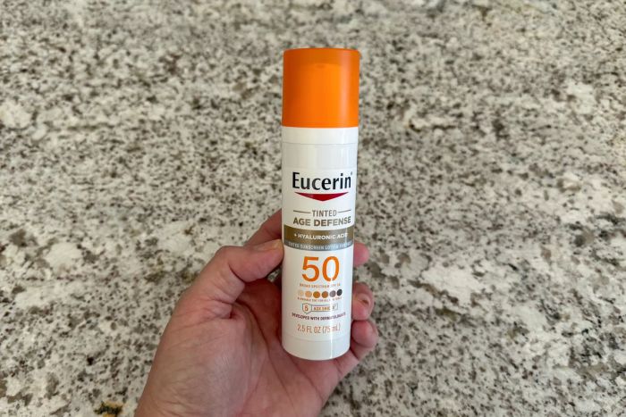 A hand holding a bottle of Eucerin Age Defense Face Sunscreen Tinted Lotion SPF 50