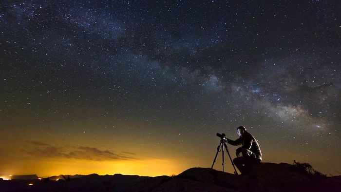 A breathtaking view of the Milky Way, with a photographer capturing the scene while seated on a rock.