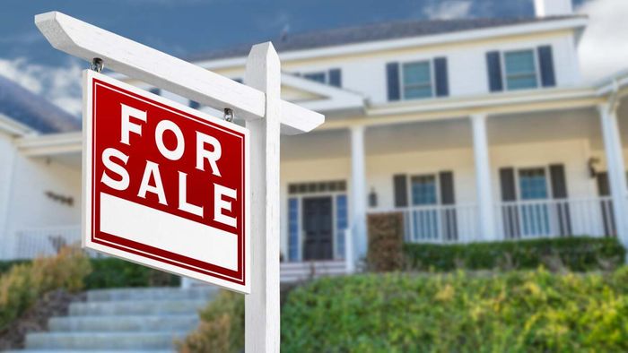 A 'For Sale' sign placed in front of a spacious white home in a suburban neighborhood.