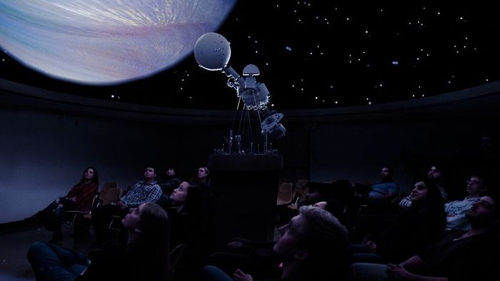 A group of individuals seated in a planetarium, observing the galaxy displayed on the dome overhead.