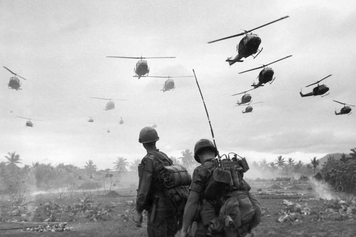 A black-and-white image captures helicopters soaring over two soldiers, their backs turned to the camera.