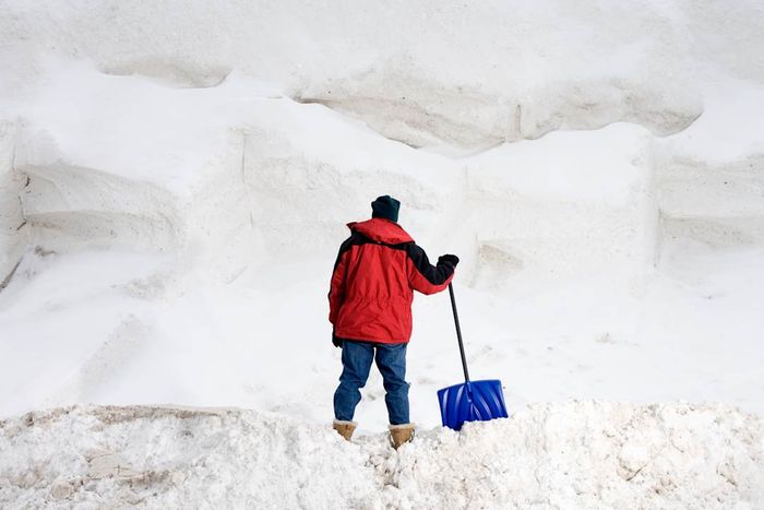 A person holding a snow shovel, looking at a large pile of snow from behind.