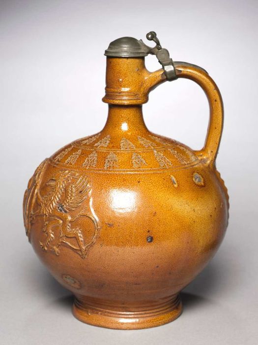 A light copper-colored, round jug with a prominent handle and a pewter lid, covering a narrow spout.