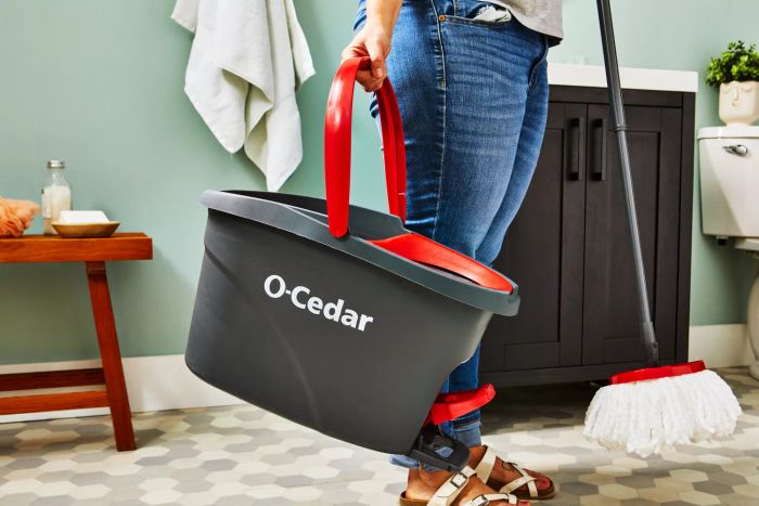 A person carrying the O-Cedar EasyWring Spin Mop and Bucket System in a bathroom with a wooden bench and sink nearby
