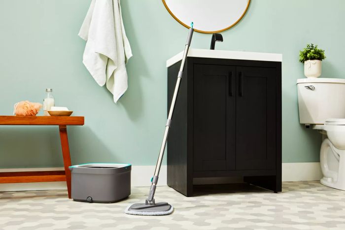 The True & Tidy TrueClean Spin Mop and Bucket System is shown leaning against a bathroom sink on tile flooring.