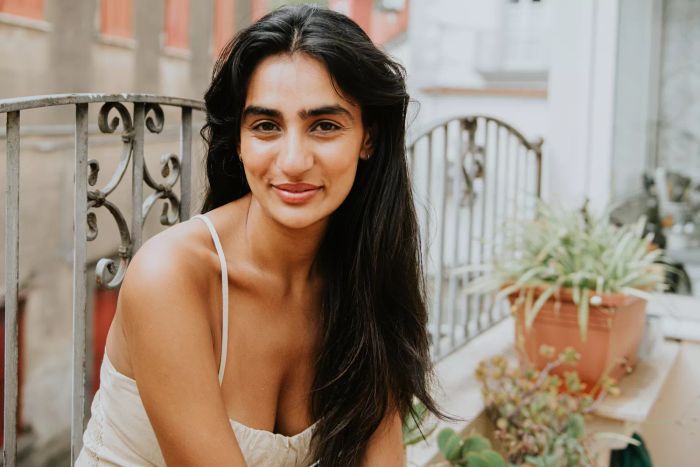 A simple portrait of a beautiful Indian woman standing on a European-style balcony, her long wavy hair cascading down. She gazes into the camera with a soft smile.