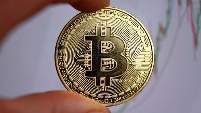 A hand grips a physical Bitcoin while a stock chart is visible in the background.