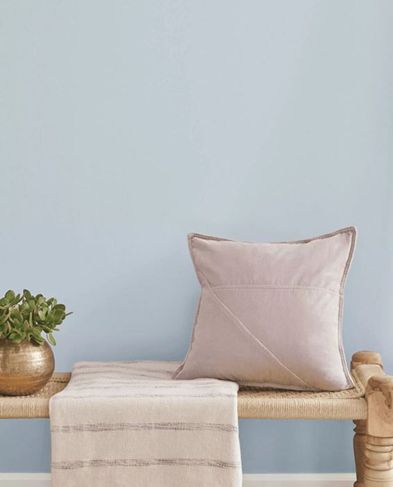 A soft light blue shade, Upward by Sherwin-Williams, adorns the wall, complemented by a woven bench, a pillow, and a blanket.