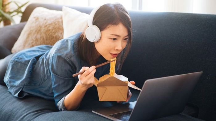 A young woman enjoying noodles while watching a movie on her laptop.