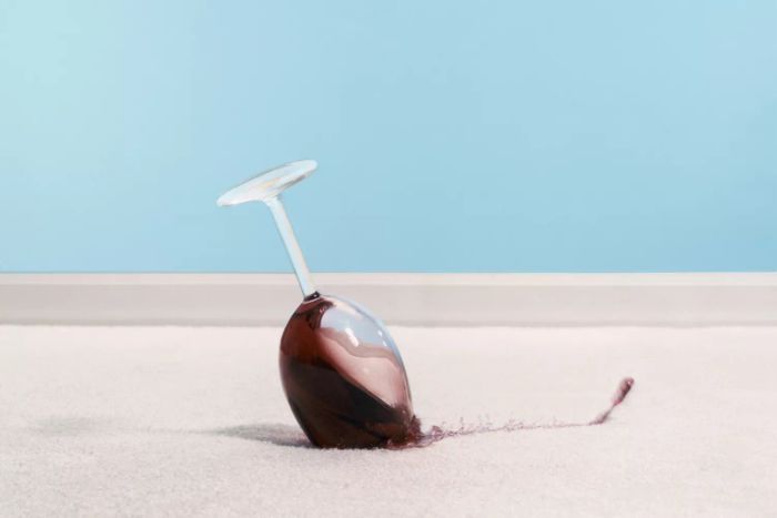 A glass of red wine spilled on a white carpet with a blue wall in the background.