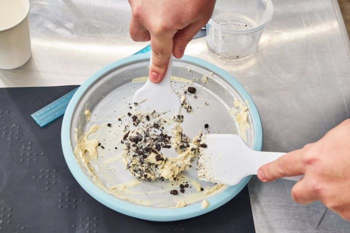Hands using white utensils to mix cookies with ice cream in the Chef'n Sweet Spot Ice Cream Maker resting on a stainless steel surface.