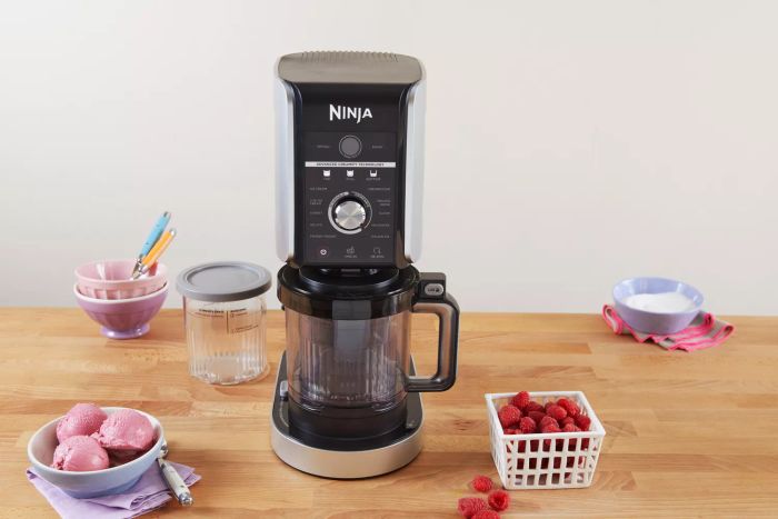 Ninja Creami Deluxe 11-in-1 XL Ice Cream Maker displayed on a wooden surface next to a bowl of ice cream and fresh raspberries