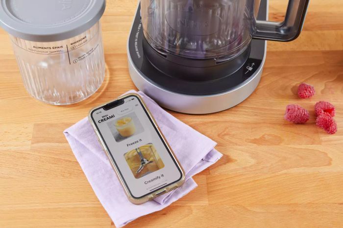Ninja Creami Deluxe 11-in-1 XL Ice Cream Maker placed on a wooden surface beside a smartphone with the Creami app open