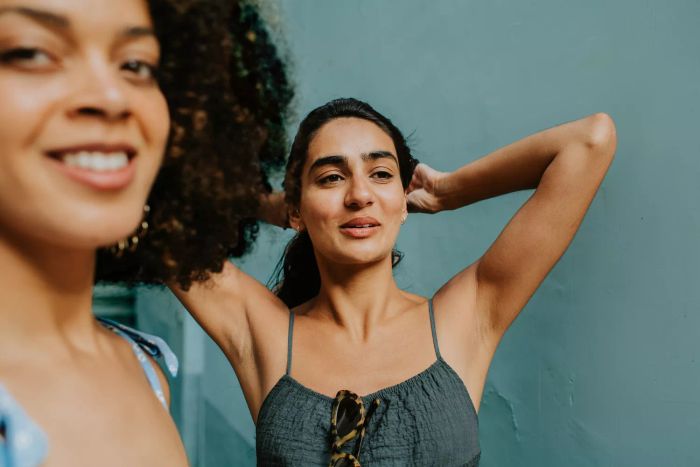 A candid photo showing two women: the one in the foreground gazes past the camera, while the other raises her arms to pull her hair back.