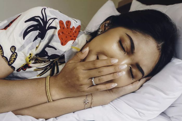 A young Asian woman, feeling drowsy, lies on her side on the bed, yawning.