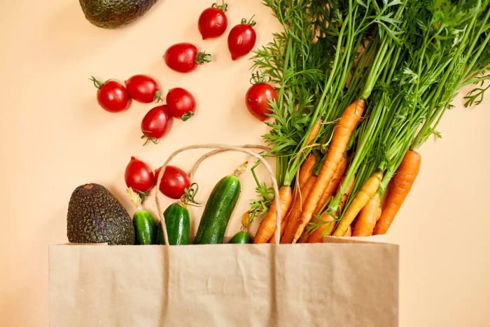 A top-down view of vegetables like carrots, cucumbers, avocados, and tomatoes packed in a paper bag on a beige background, representing healthy food and grocery shopping.