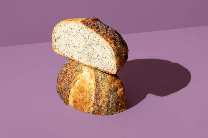 A minimalist image of freshly baked bread sprinkled with poppy seeds, placed on a purple table. The loaf of bread is sliced in half, illuminated by bright light, with a colorful backdrop.