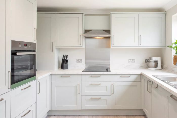 An interior shot of a freshly installed sage green and grey shaker-style kitchen, complete with stainless steel handles, an extractor fan, splashback, oven, knife block, food mixer, potted plant, recessed ceiling spotlights, and a marble-effect worktop in a home.