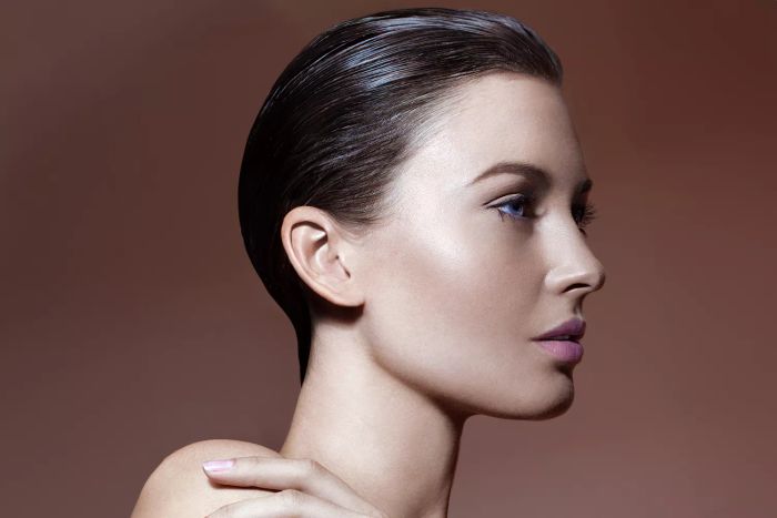 A portrait of a young, fashionable woman with wet, styled hair, sun-kissed skin, set against a rich brown background. She is in profile, her hand resting on her shoulder, with bare shoulders and neck. The look features natural makeup, striking blue eyes, flawless skin, light pink lipstick, and matching nail polish.
